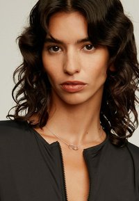 Collar de plata con acentos circulares entrelazados, llevado por una modelo con cabello rizado y oscuro. Una prenda negra con cremallera resalta el escote.