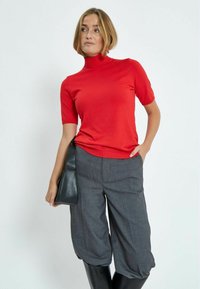 Top rojo de manga corta con cuello alto, combinado con pantalones cortos grises de corte holgado y botas negras hasta la rodilla, resaltado con un bolso negro.