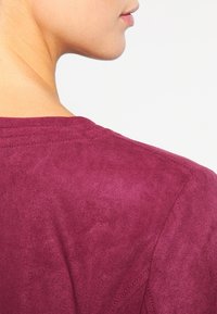 Burgundy mockasinfärgad topp med rund halsringning, synliga sömdetaljer och en mjuk textur, som visar tygets släta utseende.