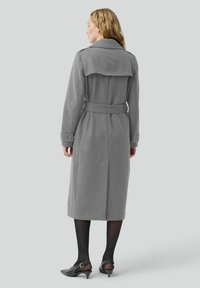 Cappotto trench grigio in lana con vita regolabile, ampio colletto e bottoni sulle maniche. Abbinato a scarpe nere con tacco e collant.