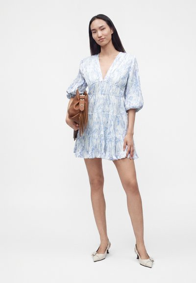 Jeune femme en robe bleu clair à motifs tenant un sac à main marron, portant des chaussures blanches à talons, debout devant un fond blanc uni.