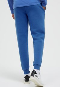 Person, die blaue Jogginghosen und weiße Sneaker mit schwarzem Fersen-Detail und "Camp David"-Branding trägt, vor weißem Hintergrund steht.