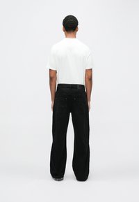 Mężczyzna stojący tyłem, ubrany w prostą białą koszulkę i luźne czarne dżinsy z tylnymi kieszeniami, na jednolitym jasnoszarym tle.