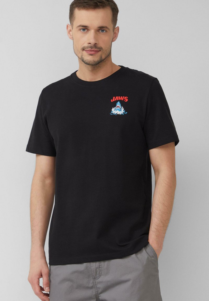 Schwarzes Baumwoll-T-Shirt mit Rundhalsausschnitt, mit rotem "JAWS"-Text und blauem Hai-Grafik auf der Brust. Kurze Ärmel, lässige Passform.