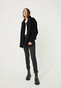 Giacca in pile nera e oversize con colletto a scialle, indossata sopra una camicia bianca. Abbinata a jeans neri attillati e stivaletti neri.