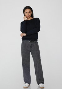 Granatowy dziany sweter z prążkowanymi mankietami, w połączeniu z szarymi wzorzystymi spodniami w szerokim kroju. Model ma na sobie białe sneakersy oraz czarny pasek.