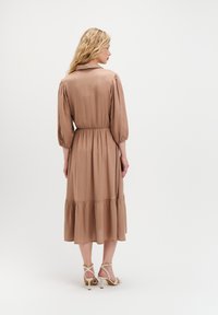 Kobieta z długimi falującymi blond włosami, ubrana w beżową midi sukienkę z bufkami i beżowe sandały na obcasie, stojąca tyłem na białym tle.