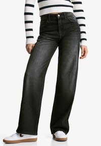 Jeans neri a gamba larga con vita alta e design a cinque tasche, abbinati a sneaker bianche. Il tessuto mostra una sottile texture denim.