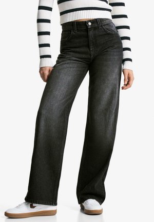 Black wide-leg jeans with a high waist and five-pocket design, paired with white sneakers. The fabric shows a subtle denim texture.