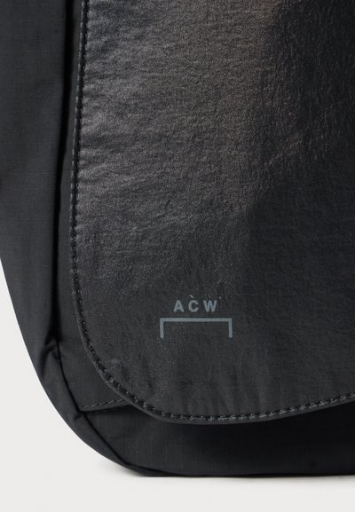 Schwarze Stofftasche mit glatter Textur, einem Klappenverschluss und einem geprägten Logo "ACW" am Boden. Verfügt über ein dezentes Streifenmuster.