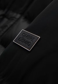 Sezione di giacca a vento nera con un tessuto testurizzato, dotata di un'etichetta nera rettangolare con la scritta "OMBRA EXIMIO" in lettere in rilievo.