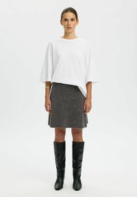 Bijela oversized majica kratkih rukava, siva vunena suknja i čizme do koljena od crne kože, stojeći na jednostavnoj bijeloj pozadini.