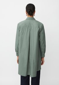 Langärmliges Shirt aus gedämpftem grünen Baumwollstoff mit einem Rückenfalten, Hemdkragen und knöpfbaren Manschetten, kombiniert mit dunkelblauen Hosen.