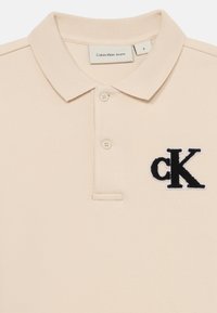 Camisa polo bege feita de tecido texturizado, apresentando um logótipo 'ck' bordado em preto no peito e uma gola clássica com dois botões.
