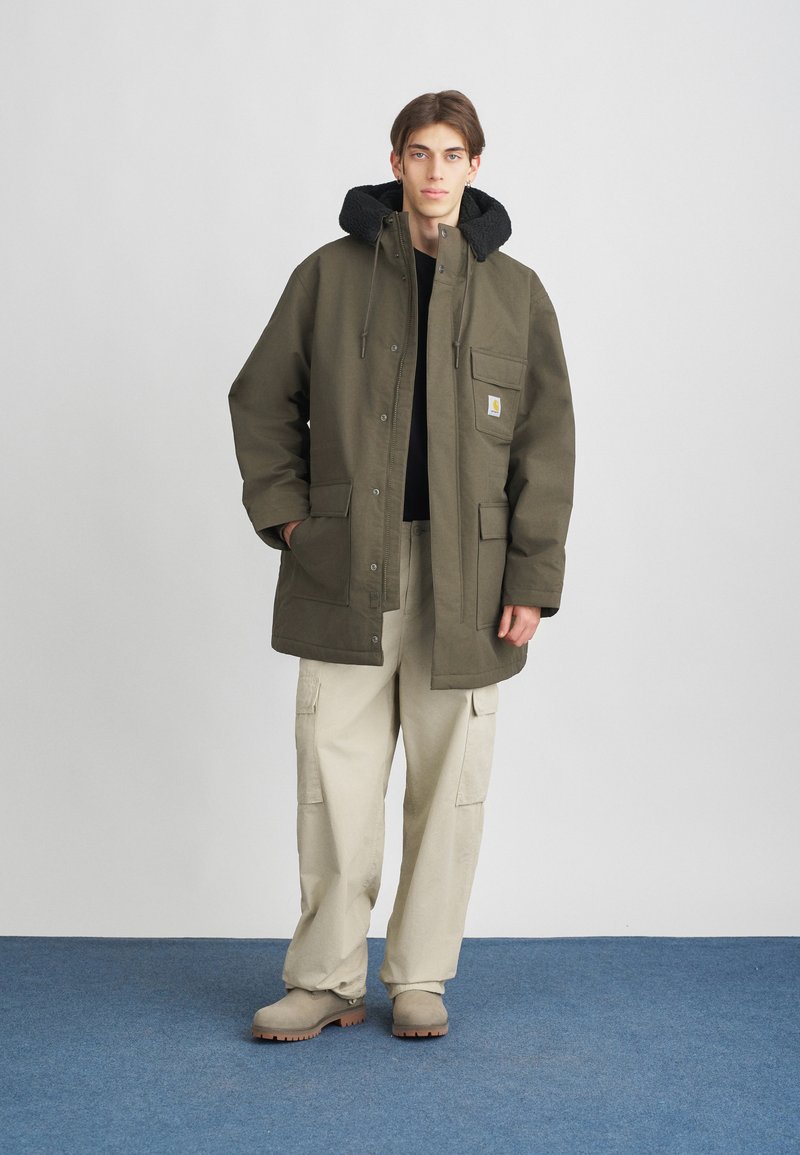 Parka verde oliva con cappuccio foderato in pile nero, numerose tasche e chiusura a zip. Indossato sopra una maglietta nera e pantaloni cargo beige chiaro.