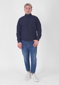 Mørkeblå zip-up jakke med høj krave, lavet af glat stof, parret med lyseblå jeans og hvide sneakers. Ingen synlige mønstre eller accenter.