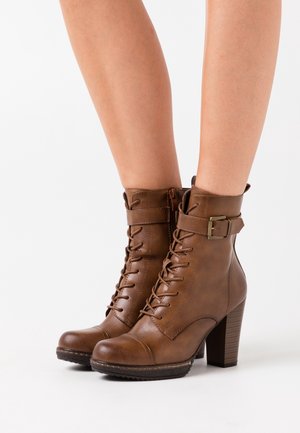 Bottes montantes en cuir marron à talons hauts avec lacets, fermeture éclair latérale et bride à boucle portées sur des jambes nues, sur fond blanc.