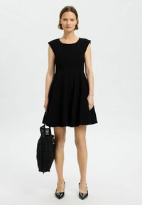 Musta pleatattu mekko pyöreällä kaula-aukolle ja hihattomilla hihoilla. Yhdistetty mustan kudotun laukun ja mustien teräväkärkisten korkokenkien kanssa.