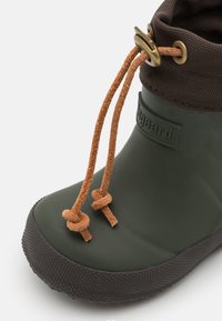 Groene rubberlaars met een gestructureerde zwarte zool, een bruine stoffen manchet en bruine lederen veter bevestigd met een antieke messing sluiting.