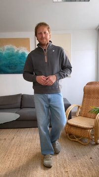Grå zip-up sweatshirt med vit och orange rand, ljusblå jeans och grå skor. Står inomhus med en soffa och en rottingstol.