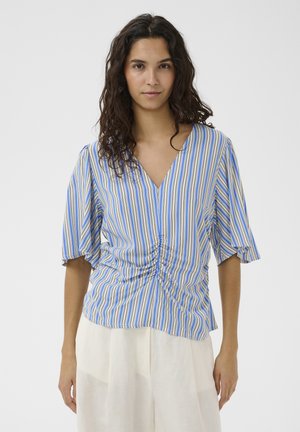 Femme aux longs cheveux bouclés et foncés portant un chemisier à rayures verticales bleu, blanc et beige avec un détail froncé et un pantalon blanc ample.