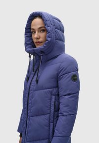 Violetti puhallettava takki korkealla kauluksella ja suurella hupulla, jossa on tikkaukset, nyöritys yksityiskohtia ja logopatchi hihassa.