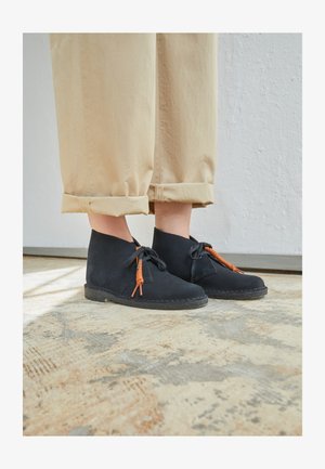 Mustat mokkanahkaiset nilkkasaappaat oransseilla nauhoilla, pyöreä kärki ja matala kumipohja; yhdistettynä käännettyihin béžinvärisiin housuihin teksturoidulla pinnalla.