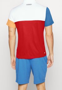 Koszulka polo z blokami kolorów w czerwonym, niebieskim, białym i pomarańczowym. Wykonana z gładkiej tkaniny, z kołnierzykiem i logo na plecach.
