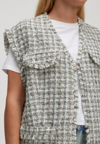 Geweven vest in lichtgrijs en beige met rafelige randen, voorzien van brede schouderflappen en een open voorkant, gelaagd over een wit T-shirt.