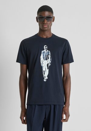 Hombre con gafas de sol oscuras, camiseta gráfica azul marino con un dibujo blanco de una figura caminando, y pantalones azul marino a juego, de pie contra un fondo gris.