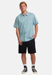 Camisa de botones de manga corta en azul claro con un patrón geométrico, combinada con pantalones cortos negros y zapatillas altas negras, con calcetines blancos.