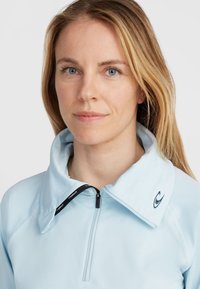 Hellblauer Reißverschlussjacke mit hochgeschlossenem Kragen, ausgestattet mit einem schwarzen Reißverschluss und einem kleinen Logo am Kragen; hergestellt aus einem glatten, atmungsaktiven Material.