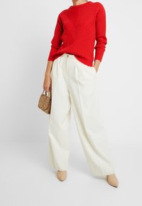 Czerwony sweter z prążkowanym wzorem w kształcie litery V, zestawiony z białymi szerokimi spodniami. Modelka trzyma małą torebkę w kolorze brązowym z motywem skóry węża. Beżowe szpilki dopełniają stylizację.