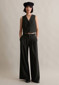 Schwarze, taillierte Weste mit V-Ausschnitt und sechs Knöpfen, kombiniert mit weiten schwarzen Hosen. Glatte Textur mit subtilen Nadelstreifen.