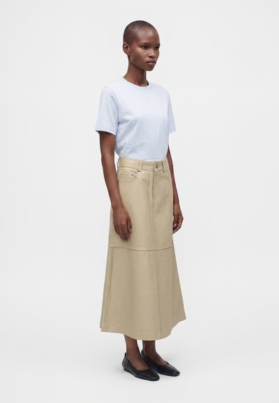 Femme portant une chemise bleu clair à manches courtes, une longue jupe beige avec des poches et des chaussures plates noires, debout devant un fond blanc uni.