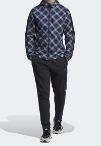 Giacca a zip a quadri neri e blu con cappuccio, maniche a contrasto e pantaloni neri abbinati con accenti blu. Design sportivo con tessuto testurizzato.