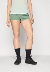 Grønne sportshorts laget av lett materiale, kombinert med svarte sokker og svarte løpesko. Shortsen har en tettsittende design.