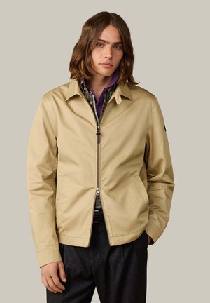 Junger Mann mit schulterlangem Haar, der eine beige Jacke mit Reißverschluss über einem karierten Hemd und einem lila Polo trägt, vor einem schlichten Hintergrund stehend.