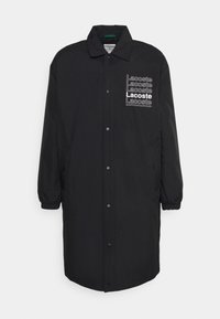 Černý dlouhý kabát vyrobený z lehkého materiálu, s uzavřením na patentky a elastickými manžetami, s několika bílými potisky loga "Lacoste".