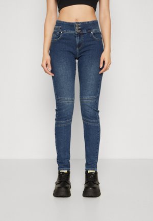 Vrouw draagt hoge taille blauwe skinny jeans met driemaal knoopsluiting en zwarte veterlaarzen, staand tegen een effen lichte achtergrond.