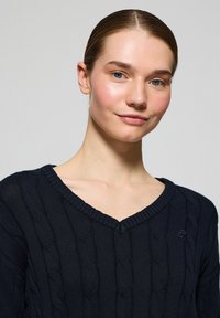 Marinblå kabelstickad tröja med V-ringning, ribbade ärmslut och liten logodetalj på bröstet. Tygtexturen är mjuk och kompakt.