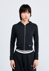 Sweatshirt preta com fecho zip e capuz, bolso frontal e logótipo da Nike em branco. Apresenta mangas ajustadas e um design desportivo. Combinado com calças de treino pretas.