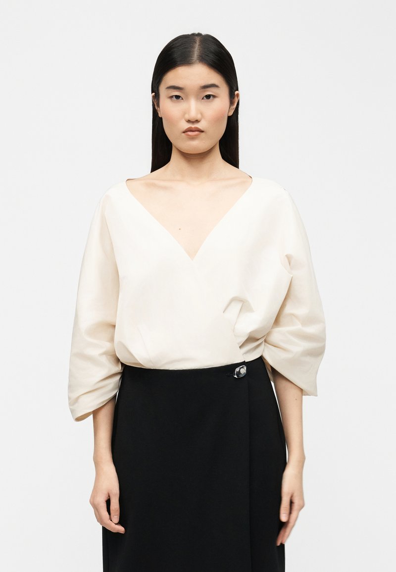 Νεαρή γυναίκα με ίσια σκούρα μαλλιά φορεί λευκό μπλουζάκι με V λαιμόκοψη και φαρδιά γραμμή, καθώς και μαύρη φούστα με ψηλή μέση, σε απλό φόντο.