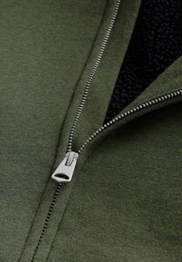Olivgr�üne Zip-Jacke mit silbernem Reißverschluss und schwarzer Fleece-Innenauskleidung. Hat eine glatte Stofftextur und saubere Nähte.