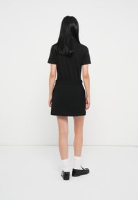 Naine, kellel on pikad mustad juuksed, kannab musta lühikeste käistega kleiti, valgeid sokke ja musti loafer-sandaale, seistes valge seina poole.