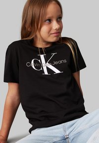 Melna kokvilnas T-kreklis ar baltu "Calvin Klein Jeans" logo; īsas piedurknes, brīvs piegriezums, gluda tekstūra, valkāts ar gaiši ziliem džinsiem.
