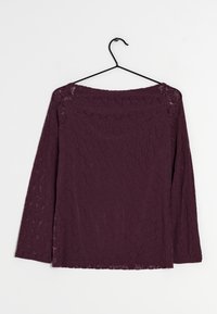 Långärmad spetsblus i djup lila. Har ett blommigt mönster och en rak halsringning. Hänger från en svart galge mot en vit bakgrund.