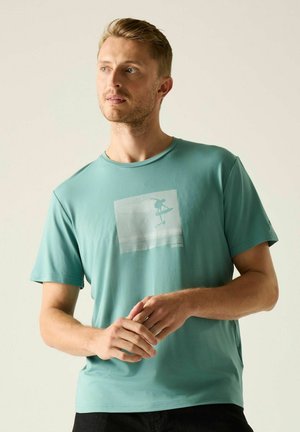 Hombre con cabello rubio corto que lleva una camiseta verde azulado con una gráfica de surfista, de pie y mirando hacia un lado contra un fondo simple.