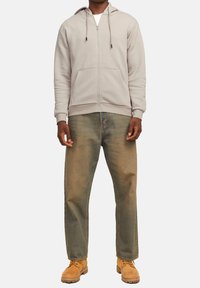 Sweat à capuche gris zippé avec cordons, poches avant, associé à un jean bleu délavé et des bottes de travail jaunes. Design simple avec une coupe décontractée.