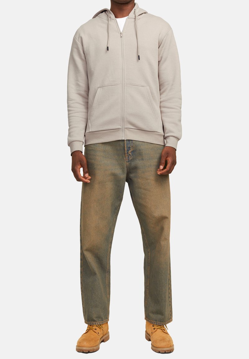 Sweat à capuche gris zippé avec cordons, poches avant, associé à un jean bleu délavé et des bottes de travail jaunes. Design simple avec une coupe décontractée.